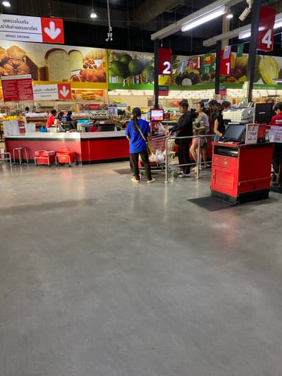 The image depicts the checkout area of a supermarket, likely located in Baan Bo Phut, Thailand, given the presence of Thai text on signage. Several people are visible, with a line of customers waiting to pay for their purchases. Two cashiers are attending to customers at separate checkout counters. The environment is a brightly lit indoor retail space with a polished concrete floor. Red and white signage is prominent, indicating departments such as "Invoice Checking" and "Goods Return." Shopping carts and baskets are also visible. The overall scene is a typical retail checkout environment during operating hours.