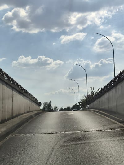 The image depicts a curved road, likely an overpass or a section of highway, under a bright, cloudy sky. The road surface is asphalt, showing signs of wear and a visible seam running down its center. On the left side of the road, a concrete barrier with a metal railing forms the edge. Several modern streetlights with curved arms are spaced along this barrier, their lamps pointing downwards. Beyond the barrier, the sky is filled with white and gray cumulus clouds, suggesting daytime with partly cloudy conditions. Trees line the distant edge of the overpass, with some foliage visible between the streetlights. A triangular warning sign with a red border is partially visible among the trees. The overall impression is one of a mundane, everyday scene in an outdoor setting, possibly during the day. There is no visible text in the image. The orientation of the image suggests it was taken from a low angle, looking up at the sky and along the road.