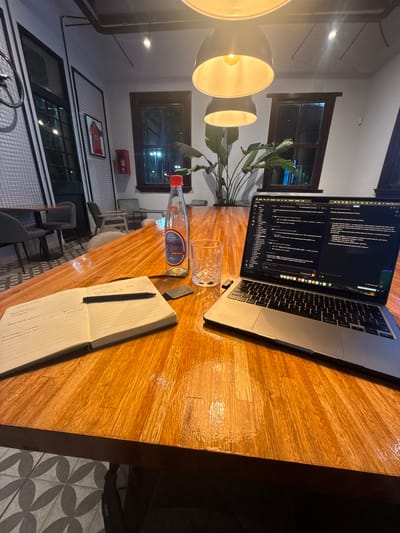 The scene is a workspace on a wooden table in a café or restaurant in Avellaneda, Argentina. A laptop displays lines of code on its screen, placed next to a half-full glass of water and a bottle of “Bermuda” brand water. A notebook lies open with notes written on its pages next to a pen. The room is lit by a modern, multi-bulb pendant lamp. Through the windows, we can see the exterior setting. There is a framed poster on the wall and a tiled floor with a geometric pattern. Several chairs are visible, indicating that this is a seating area for customers.