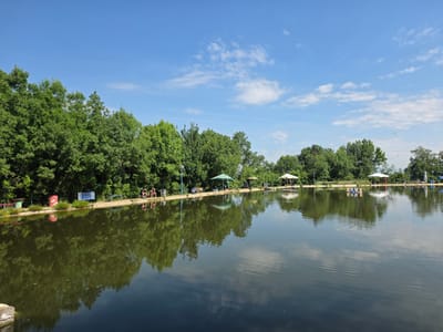 The image depicts a serene lake scene under a bright blue sky with scattered white clouds. Along the edge of the lake, a line of lush green trees forms a dense backdrop. Several individuals are visible on the shore, some seated on benches, appearing to be engaged in recreational activities, possibly fishing. Umbrellas are set up in various locations along the bank, providing shade. A few small structures and objects, such as bins and a sign, are also present. The calm water of the lake reflects the trees and the sky, creating a mirror-like effect. The overall atmosphere is one of a peaceful outdoor environment. The location is identified as a lake in the city of German, Bulgaria.