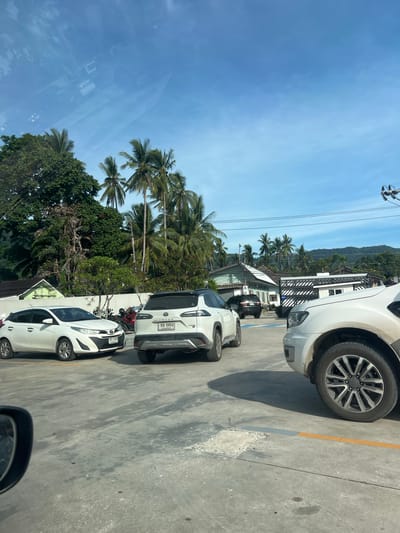 In Baan Lamai, Thailand, a daytime outdoor scene captures a parking area under a clear blue sky with scattered clouds. Several white cars are parked on a concrete surface. A white hatchback is on the left, a white SUV-style vehicle is in the center, and a portion of another white vehicle is visible on the right. Behind the vehicles, buildings with white walls and dark roofs are situated among lush green trees, including numerous palm trees. A mountain range is visible in the distance. The atmosphere suggests a warm and sunny environment.
