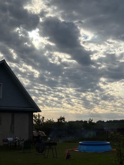 In an outdoor setting, possibly in Мётчанскі сельскі Савет, Belarus, a grey and blue building with a prominent gable end is partially visible on the left. In front of the building, a small dining set with a table and chairs is arranged on a grassy lawn. Nearby, a charcoal grill is set up, with a small fire burning and emitting smoke. To the right of the grill, a blue inflatable pool sits on a striped mat. The background features a dense line of trees and a wooden fence. The sky above is dramatic, filled with a textured pattern of grey and white clouds, with sunlight breaking through in several areas. The overall scene suggests a relaxed, possibly evening, gathering in a rural or suburban environment.