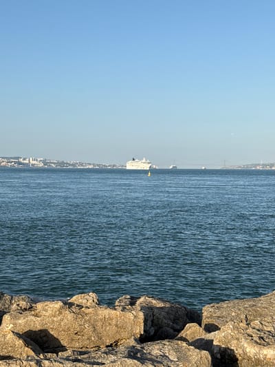 A wide-angle, eye-level, vertical shot captures a bright, sunny day with a clear, pale blue sky. The foreground is dominated by large, light-brown rocks with deep shadows, suggesting a rocky shoreline. Beyond the rocks, a vast expanse of dark blue, slightly rippled water stretches to the horizon. In the mid-ground, a white cruise ship is visible on the water, and further in the distance, a cityscape with numerous buildings lines the shore. A large red suspension bridge spans the water in the far distance. The scene appears to be the Tagus River, likely near Oeiras, Portugal, given the presence of the suspension bridge, which resembles the 25 de Abril Bridge. No people are clearly visible, and the focus is on the natural and man-made elements of the waterfront.