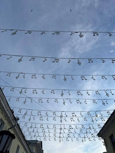 The image presents a daytime scene looking upwards towards a partly cloudy sky, featuring an extensive overhead display of decorative lights shaped like musical notes. Numerous rows of these light-colored decorations, primarily depicting treble clefs and eighth notes, are suspended by wires across the view.
The sky, a bright blue with scattered white clouds, fills most of the frame. Two small, dark birds are visible in flight against the sky on the left side, one more prominent than the other.
On the right side, the upper section of a building is visible, including a roofline with an overhang, a gutter, and a section of a wall, possibly light-colored. The decorations appear to extend from fixtures associated with this building.
The scene suggests a festive or themed public area in Заволжье, Russia, adorned with musical motifs. The clear daylight indicates good weather. No visible text is present in the image.