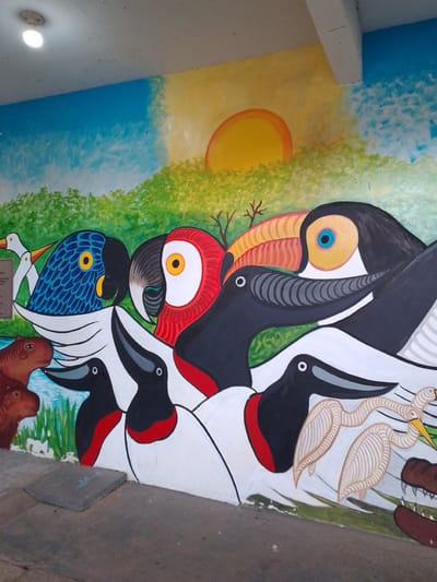 This image depicts a colorful mural painted on a wall in Cáceres, Brazil, featuring various animals. In the upper portion of the mural, a bright yellow and orange sun is setting over a landscape with green trees and a blue sky. A prominent feature of the mural is a group of stylized birds. Among them are a blue parrot, a red and white macaw, and a toucan with a large, multi-colored beak. Several other birds with black and white plumage and red markings on their chests are also visible, along with two slender, light-brown birds that appear to be cranes or herons. In the lower left of the mural, two capybaras are depicted near the water. To the right, the jaws of a crocodile are visible. The mural appears to be located outdoors, possibly along a sidewalk or walkway, given the concrete ground visible in the foreground and a square concrete slab. The overall style of the artwork is vibrant and somewhat simplified, characteristic of folk art.
