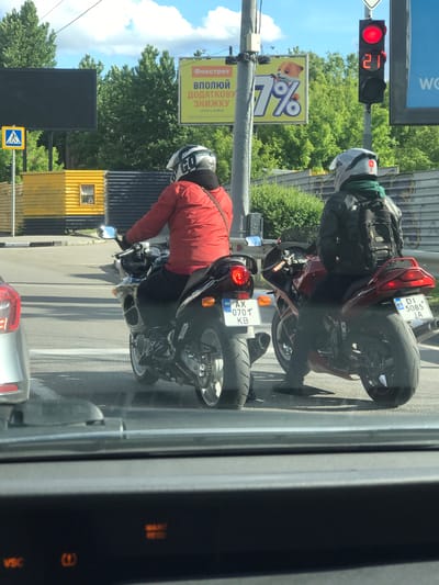 The image shows two motorcyclists stopped at a traffic light. The first motorcyclist is wearing a red jacket and white helmet. The second motorcyclist is wearing a black jacket and white helmet. Both motorcycles have Ukrainian license plates. The traffic light is red and shows 21 seconds remaining. Behind the motorcyclists are trees and buildings, including a billboard for "Fokstrot" and a yellow and black building. The photo was taken from inside a vehicle, showing part of the dashboard. It is not possible to determine the location of the photo within the city of Lima, Peru as this information is not apparent in the image.