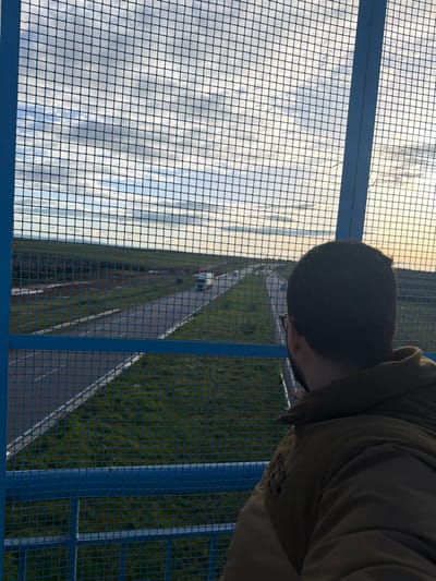An elevated view from a blue metal safety barrier or pedestrian bridge in Ait Boubidmane, Morocco, shows a person observing a dual-carriageway road. The individual, seen from behind in the lower right quadrant, has short dark hair and wears glasses and an olive-green jacket, looking towards the distant horizon. The blue metal grid of the barrier occupies the foreground. Below, a two-lane road in each direction features white line markings, flanked by green grass. A white semi-truck travels away from the viewer on the left side of the road, with other smaller vehicles visible further down. The road extends through a flat, sparsely vegetated landscape under a sky with scattered white and grey clouds, illuminated by soft, possibly early morning or late afternoon light.