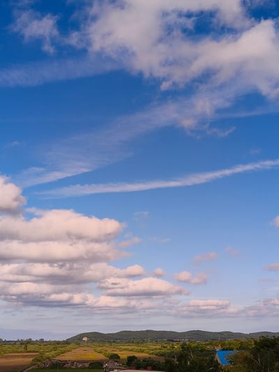 This media file captures a wide-angle view of a landscape under a vast, cloudy sky. The upper portion of the image is dominated by the blue sky, with scattered white and pink-tinged clouds. Some of the clouds are wispy and stretched out, suggesting movement or high altitude, while others are more voluminous and soft-edged.

The lower half of the image depicts a rural or semi-rural landscape. Rolling green hills, covered in dense vegetation and trees, form the backdrop. In the middle ground, there are fields, some appearing cultivated and others more overgrown with bushes and trees. A few scattered buildings are visible, including a house on top of one of the hills and a building with a distinctive blue roof in the foreground on the right. The overall impression is one of a serene, natural setting.

The time of day appears to be late afternoon or early evening, given the soft lighting and the warm hues present in some of the clouds. The weather seems pleasant, with a mix of sun and clouds creating a dynamic sky. There are no visible people or animals. The location is contextually identified as Reda, Poland. The image conveys a sense of peace and natural beauty, with the expansive sky and the verdant landscape offering a tranquil scene. There is no visible text in the image.