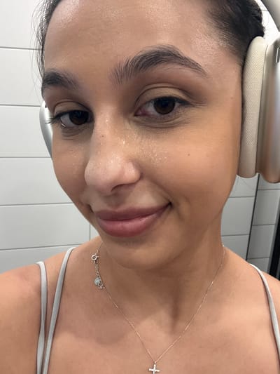 The image shows a close-up, eye-level, medium shot of a young woman with dark hair, wearing a light-colored tank top and a silver necklace with a cross pendant. She is also wearing silver and beige headphones. Her face is slightly sweaty, and she is smiling. The background appears to be a tiled wall, suggesting an indoor location like a gym or bathroom. There is no visible indication of the city of Mönchengladbach, Germany.