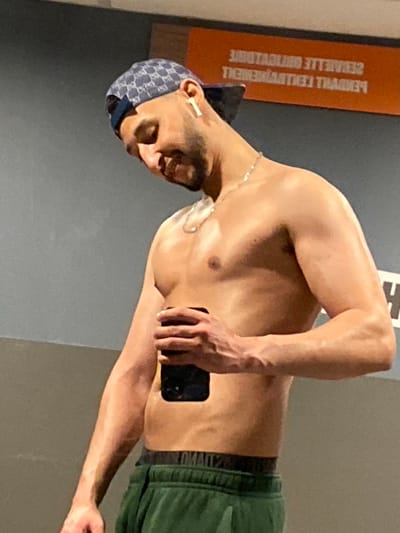A shirtless male individual is depicted from the mid-chest upwards, observed in what appears to be a mirror reflection. He faces slightly right, his head tilted downwards, exhibiting a subtle smile. He wears a blue patterned baseball cap backward, a white wireless earbud in his right ear, and a thin silver chain necklace. His physique is muscular, and his skin appears moist. He holds a black smartphone vertically in his left hand, obscuring a portion of his lower torso. Green athletic pants with a black elastic waistband are visible below his waist. The background consists of a grey wall and an orange panel bearing inverted white French text that reads "SERVIETTE OBLIGATOIRE" (Towel obligatory) and "ENTRAINEMENT" (Training). This scene is consistent with an indoor fitness facility, specifically located in Hœnheim, France, based on the linguistic context of the background text.