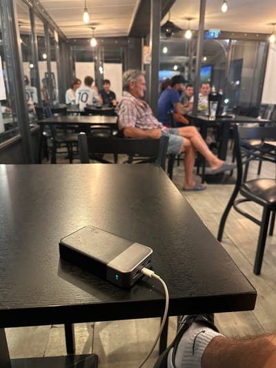 This is a close-up, eye-level shot of a black wooden table at what appears to be an indoor cafe or restaurant, with a power bank plugged into a device, partially obscuring the view of the rest of the establishment and its patrons. The power bank is metallic grey and black, with a white cable connected to it, the other end of which is not visible. A green light indicates it is active. The table surface exhibits a visible wood grain. In the background, a man in a plaid shirt and denim shorts is seated at a table, with his legs crossed and wearing blue slippers. Other patrons are visible at tables behind him. The environment suggests a casual dining or gathering space. The location of Anduze, France is not discernible from the image itself.