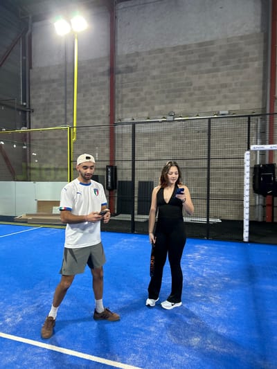 Two individuals, a man and a woman, are captured posing on an indoor padel court. The bright blue court surface features white boundary lines, with tall black mesh fencing and gray concrete block walls enclosing the playing area. Overhead, artificial lighting illuminates the scene.
The woman stands on the right, wearing a black tank top and black fitted leggings, paired with white athletic shoes. She holds a dark smartphone in her right hand, positioned near her chest. A padel racket with an orange and black pattern is partially visible by her left leg. She has long brown hair.
On the left, the man is dressed in a white short-sleeved jersey featuring the Italian flag emblem and the word "ITALIA" on the left chest. He wears gray shorts, white socks, and brown athletic shoes. A white baseball cap is worn backward on his head. He is also holding a dark smartphone in his left hand.
Both subjects appear to be standing still, possibly for a photograph, rather than actively playing. In the background, part of the padel net system is visible, with white posts displaying the word "PADEL" repeatedly. A black sign further in the background shows partially legible text "WWW.SOCCERBLU...". The setting is an indoor sports facility, specifically a padel court located in Eckbolsheim, France. The presence of artificial lights indicates an indoor setting where the time of day is not a factor.