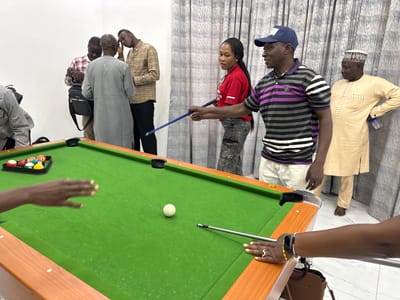 A group of people are gathered around a pool table in an indoor setting. The room appears to be a recreation area, possibly a home or a community center, with a light-colored wall and patterned curtains in the background.

On the pool table, a rack of billiard balls is set up, and a white cue ball is positioned in the center. Several individuals are interacting with the table and each other. One man, wearing a purple and black striped polo shirt and a blue baseball cap, is holding a pool cue, seemingly preparing to take a shot. A woman in a red shirt and jeans stands nearby, watching.

Other individuals are visible in the background. One man in a tan and brown plaid shirt is looking down at something in his hands, while another man in a grey traditional outfit stands beside him. A man in a light yellow traditional outfit with a white cap is standing to the right, observing the scene. The presence of a pool table and the casual attire suggest a leisurely gathering or a friendly game.

The lighting appears to be artificial, indicating that the event is taking place indoors, and the time of day is not determinable from the image. The overall atmosphere seems relaxed and social. No visible text is present in the image. The location context provided is Unguwar Sarki, Nigeria.