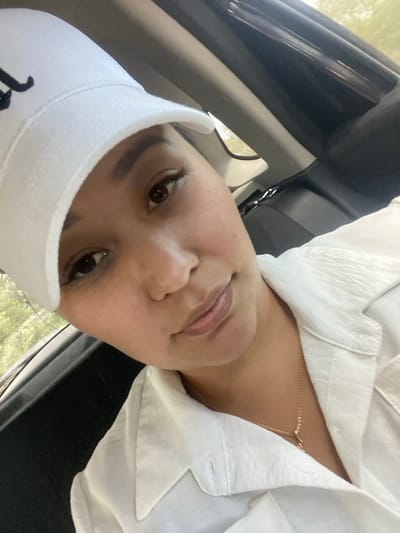 A woman is captured in a close-up, angled selfie, her face tilted towards the camera. She wears a white baseball cap with a black logo and a white collared shirt, which is partially unbuttoned, revealing a delicate gold necklace. The background appears to be the interior of a vehicle, suggested by the dark, contoured surfaces that resemble car upholstery and window trim. Sunlight filters in, creating a softly lit environment. The woman's gaze is directed slightly off-camera, and her expression is neutral. The image is devoid of any identifiable landmarks within Akyar, Russia.