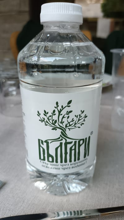 The image shows a clear plastic bottle of water. The bottle has a white cap and a label with a green tree logo and text in Bulgarian. The text on the label reads "БЪЛГАРИ" and below that "Свързани чрез корените, освежени чрез водата!". The bottle is sitting on a light-colored, textured surface, likely a tablecloth, with a metallic object partially visible in the foreground. In the background, blurry figures and objects suggest an indoor or semi-outdoor setting. There is no specific indication of the city of Razlog, Bulgaria within the image itself.