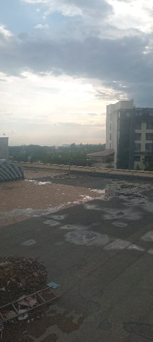 The image depicts an elevated outdoor scene in Kisumu, Kenya, under an overcast sky with bright light on the horizon. The immediate foreground features a dark, uneven ground surface, likely concrete or asphalt, extensively covered with shallow water puddles reflecting the sky. To the lower left, a pile of dark, irregular debris, possibly composed of soil and broken construction materials, is present. A metallic ladder lies horizontally on the ground adjacent to this debris. Further left, a lighter brown, sandy-textured area also exhibits wet patches.

A low, light-colored concrete parapet wall spans the midground. Beyond this wall, an urban landscape unfolds, characterized by green vegetation and various structures, including a building with a reddish-brown tiled roof. Dominating the right side of the frame is a multi-story modern building, distinguished by lighter upper sections and a darker, gridded facade on its lower levels. The distant background reveals a hazy horizon line, with a visible body of water, consistent with Lake Victoria, situated behind a line of trees. The sky overhead is predominantly gray and overcast, with several small dark specks, interpreted as birds, scattered across the brighter segment near the horizon. No human figures or discernible actions are present in the scene.