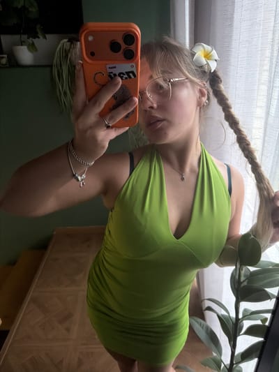 The image shows a young woman with blonde hair in a braid, wearing a green dress and glasses. She is holding an orange phone with a leopard print case, taking a selfie. A white flower with a yellow center is tucked into her braid. She is also wearing a silver necklace and bracelets. The background suggests an indoor setting with a window covered by a sheer curtain and a houseplant to the right of the frame. The lighting indicates it might be daytime. The overall mood appears casual and relaxed. There is visible text on a card inside the phone case, appearing to be "USD" and possibly a logo or image of a leopard.