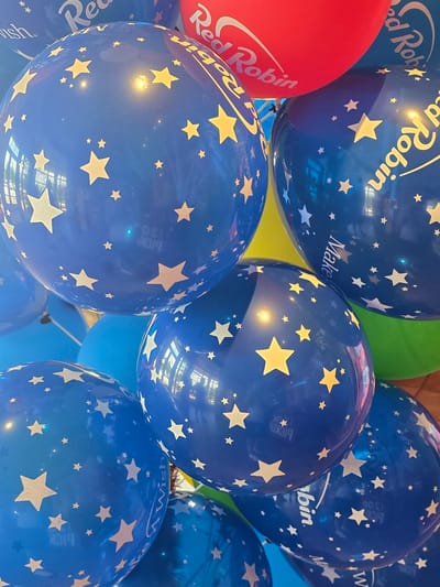 This is a close-up, eye-level shot of a cluster of blue balloons decorated with white and yellow stars. Some of the balloons also feature text, including "Red Robin" in white script and "Make a Wish" in white. A red balloon with "Red Robin" is partially visible at the top of the frame. The balloons are arranged in a dense grouping, obscuring most of the background. A sliver of green and yellow is visible behind the blue balloons, suggesting other balloons or decorations. The lighting creates reflections on the glossy surface of the balloons, highlighting the star patterns and text. There are no people clearly visible in the image, though faint reflections suggest there might be some out of frame. The scene appears to be indoors, possibly at a party or event. The city of Cumming, United States of America, is not depicted or suggested by the visual elements in this image.