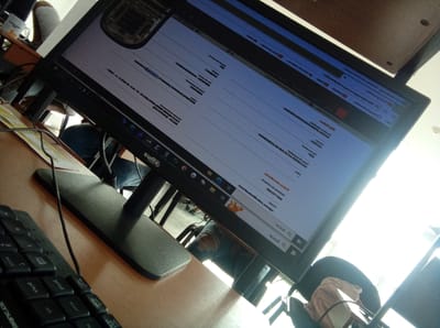 The image shows a workstation setup, possibly in an office or study. A black computer monitor dominates the scene, displaying a document or webpage filled with text. To the left, a black keyboard is partially visible on a wooden desk. Behind the monitor, a person is seated in a chair, wearing jeans. The overall environment appears to be indoors, with natural light coming from a window. The city of Parroquia Guatire, Venezuela cannot be determined from this image.