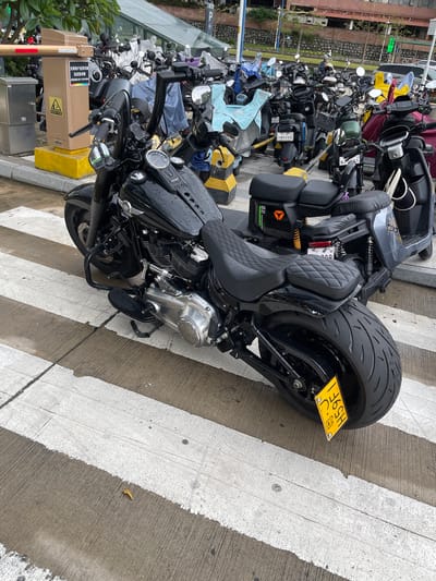 This media file shows a collection of parked motorcycles and scooters on a street, likely in Panyu District, China. The scene is an outdoor setting, with a paved surface marked with white lines, suggesting a designated parking area or a crosswalk. The time of day appears to be overcast, with the lighting suggesting it's either early morning or late afternoon. The weather seems to be damp, as indicated by water droplets visible on the motorcycles.
The main subjects are various types of motorcycles and scooters, predominantly black, with a prominent custom motorcycle in the foreground featuring a large rear tire and a yellow license plate. Several other smaller scooters and motorcycles are parked behind it. The surrounding environment includes a building with a stone facade and some greenery, possibly trees or bushes. There is also a parking barrier and an electronic kiosk visible, along with signage.
Visible text on the signage includes: "仅限商户送货车辆 及应急车辆" which translates to "Only for merchant delivery vehicles and emergency vehicles." Below this, there's a service phone number: "服务:020-31038666". Other visible text on license plates includes "HM6998", "61827", and "H59F1". There's also "02" visible on a part of one of the scooters, and "YEO" visible on a logo.
The overall impression is of a typical urban scene with a focus on parked vehicles in a functional area. There are no people directly visible in the main frame of the image. The image is captured from a slightly elevated, tilted perspective.