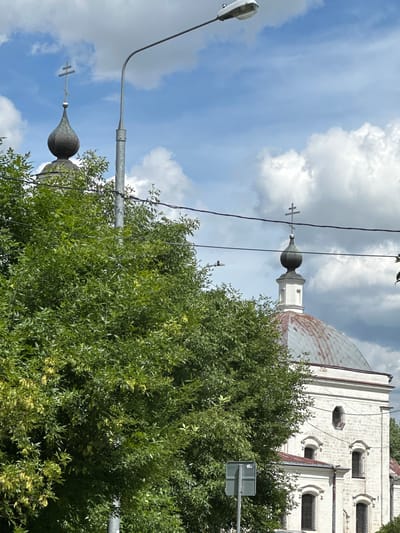 A close-up, low-angle outdoor shot captures a portion of a white stone church building with a domed roof, featuring two onion domes topped with Orthodox crosses, situated in the village of Ерино, Russia. The church's facade shows multiple arched windows. A large, dense green tree with some yellow seed pods dominates the left side of the frame, obscuring part of the church and a gray utility pole with electrical wires running across the scene. A street light on a pole is visible in the upper portion of the image against a sky filled with white, puffy clouds. The overall scene suggests a sunny day.