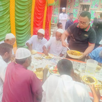 The scene, set in Lalmohan, Bangladesh, depicts a communal meal involving approximately 10-12 men in an outdoor or semi-outdoor setting. In the foreground and midground, at least seven men, predominantly elderly with beards, are seated around a table actively consuming food from plates. Most are dressed in white panjabis and white prayer caps; one seated man wears a maroon garment. The table is covered with newspaper sheets and displays plates containing rice, meat dishes, and a side salad of sliced vegetables. Clear water glasses and a plastic water jug are also on the table. A man wearing a black polo shirt with a "GUCCI" logo and eyeglasses stands, using a spoon to serve a meat dish from a white bowl onto a plate for one of the seated individuals. In the background, additional men are visible standing, including one in a white panjabi and topi with crossed arms. The environment features decorative fabric backdrops with vertical stripes in green, yellow, and red, and part of a building constructed with corrugated tin walls, including a window secured with metal bars.