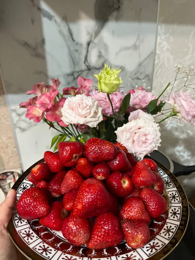 The image displays an indoor scene focused on a plate of fresh strawberries held by a human hand, set against a background of a floral arrangement and a tiled wall.

In the foreground, a round, shallow ceramic plate is filled with numerous ripe, red strawberries. Many of the strawberries exhibit a glistening surface, indicating moisture, and some retain small green caps. The plate has a distinctive decorative rim featuring a repeating pattern of white square sections containing a dark, stylized star or snowflake motif, all bordered by a metallic gold band. The center of the plate is a solid dark reddish-brown color. A fair-skinned hand is visible on the lower-left side, holding the plate by its rim.

Behind the plate, a bouquet of fresh flowers is arranged. The bouquet primarily features light pink and white Lisianthus flowers with ruffled petals, several smaller pink Alstroemeria blooms, and a single pale green/yellow Lisianthus bud or flower. Delicate white Gypsophila (Baby's Breath) is also interspersed throughout the arrangement.

The background consists of light-colored, vertically oriented wall tiles with gray marble-like veining, consistent with a kitchen or bathroom backsplash. A prominent vertical grout line is visible towards the center of the background. Reflections on the tiles suggest an overhead light source. On the far right, a dark, blurred object, possibly an appliance or countertop edge, is partially visible. On the far left, a faint, blurred metallic object is discernible.

There are no discernible details within the image to confirm the location as the city of Kaspiysk, Russia.
