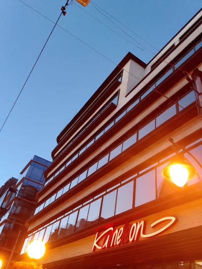 This low-angle shot captures a modern building with a curved facade against a clear blue sky. The building features multiple levels of windows with dark frames, reflecting the sky and surrounding lights. A neon sign with white text that reads "Kane on" is prominently displayed on the lower level, illuminated and glowing. Several streetlights, one with a bright yellow-orange glow, are visible, suggesting the image was taken in the evening or at dusk. The scene appears to be an urban environment. The overall mood is contemporary and urban.
