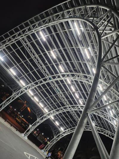 The image depicts a large, modern, covered walkway structure at night in Thủ Đức, Vietnam. The walkway's roof is supported by a complex steel truss system, illuminated by numerous bright, linear LED lights. The perspective is from below, looking upwards at the intricate metalwork and the rows of lights against a dark sky.
In the background, a street or parking area is visible. A yellow bus with visible text, possibly advertising, is parked on the left. Several palm trees with distinctive fronds are also present, suggesting a tropical or subtropical environment. The ground is paved, with white markings indicating a road or parking area. A single person, dressed in a blue shirt and dark pants, is visible on the paved area in the lower right corner, walking away from the camera. The overall scene suggests an outdoor public space, possibly a bus station, market, or transportation hub, that is operational and well-lit at night. The lighting creates a bright, almost futuristic atmosphere within the covered area, contrasting with the darkness of the night sky. The weather appears clear.