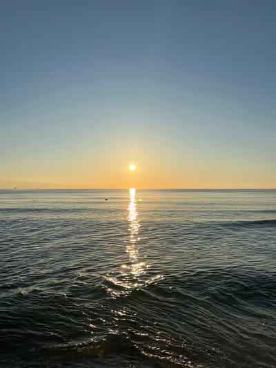 A sunset over the Baltic Sea is depicted in this image, taken from the coast of Baltiysk, Russia. The sky is a clear, soft blue, transitioning to a pale yellow near the horizon where the sun is setting. The sun itself is a bright, radiant orb, casting a shimmering reflection on the water's surface. Gentle waves roll towards the shore, their dark surfaces catching the golden light, creating streaks of sparkle. In the distance, the faint silhouettes of a few small boats are visible on the water. The overall scene is one of tranquility and natural beauty.