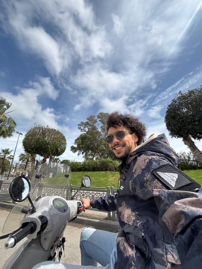 A male individual is seated on a light grey scooter under a blue sky with scattered white clouds. He possesses dark curly hair, a short beard, and wears round sunglasses, a multi-patterned hooded jacket with a white triangular patch on the left sleeve, and light blue ripped jeans. The man is smiling and looking towards the camera, with his right hand positioned on the scooter's handlebar. The scooter features a clear windscreen, visible rearview mirrors, and a digital dashboard, with keys in the ignition. The background includes a grassy area with various trees, including neatly trimmed spherical bushes, bordered by a decorative metal fence with geometric patterns. In the distance to the left, palm trees, streetlights, and buildings are visible. The scene is located in a park or promenade area within the city of Tanger, Morocco.