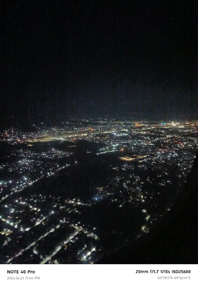 An aerial nighttime photograph captures the sprawling urban landscape of Dushanbe, Tajikistan. The city is illuminated by a dense network of artificial lights, creating a mesmerizing pattern of light and shadow. Individual buildings and streets are discernible, though details are softened by the distance and the low-light conditions. In the foreground, what appears to be a residential area with smaller, densely packed structures is visible, with streetlights casting pools of illumination. Further into the background, larger commercial or industrial areas can be identified by clusters of brighter lights. The overall impression is of a vibrant, active city extending into the darkness. The photograph includes metadata indicating it was taken with a "NOTE 40 Pro" camera on "2026.06.21 11:44 PM" with camera settings "23mm f/1.7 1/13s ISO25600" and GPS coordinates "40°28'2"N 49°56'41"E".