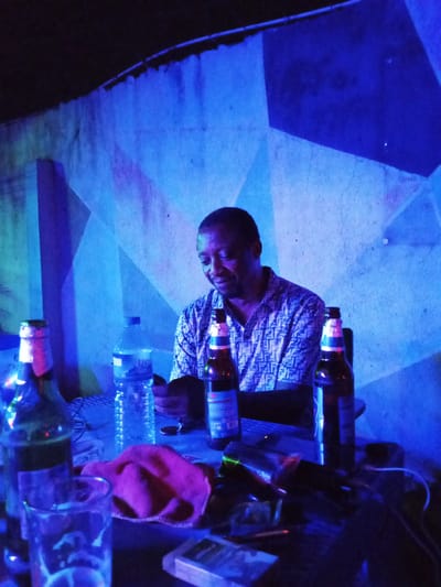 A man sits at a table in what appears to be an outdoor establishment with blue lighting and a geometrically patterned wall in Yelwa, Nigeria. He is wearing a patterned short-sleeved shirt and looking down at a phone in his hands. On the table in front of him are several bottles of drink, including what look like beer bottles, and a bottle of water. There is also a pink cloth and a pack of cigarettes. The overall ambiance suggests a casual, perhaps nocturnal, setting.