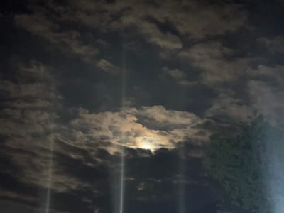 The image shows a nighttime sky filled with dramatic clouds illuminated by the moon. Several vertical light beams, possibly from searchlights or reflections, pierce through the clouds. A tree with dark green foliage is visible on the right side of the frame, partially obscuring the sky. The scene appears to be an outdoor view in Kapan, Armenia.