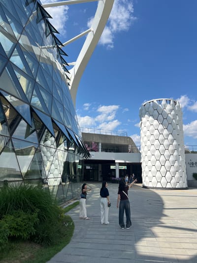 The image captures a sunny, clear day at a modern architectural complex. The scene features two prominent buildings, three individuals, and paved walkways amidst some greenery.
On the left, a large, contemporary building features an intricate glass-paneled facade, appearing as a geodesic or faceted dome structure. Its surface is composed of numerous triangular glass sections that reflect the bright blue sky and scattered white clouds. White, curving structural elements arch gracefully over and around this glass edifice.
To the right, a cylindrical or partially cylindrical building stands tall, distinguished by its striking white exterior. This facade is covered in a uniform pattern of raised hexagonal elements, creating a three-dimensional, honeycomb-like texture. Visible text on this building reads "식물문화" (Plant Culture) and "Botanic Cafe," suggesting its function.
On the paved area in the foreground and middle ground, three individuals are present. They are casually dressed and appear to be observing the architecture, with their gazes directed upwards towards the buildings. One person, wearing a dark top and dark pants, has their arms slightly raised, seemingly gesturing or taking a photo. Another person in a dark top and light pants stands nearby, also looking up. The third individual, further left, is similarly engaged.
The time of day is bright daylight, evidenced by the clear blue sky, scattered white cumulus clouds, and strong shadows cast by the buildings and people on the paved ground. Lush green bushes and grass are visible in the foreground and along the base of the buildings, adding a natural element to the structured environment. The overall impression is one of modern design and an open, inviting public space.