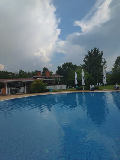 The image captures a scenic view of an outdoor swimming pool area, likely in Yagoda, Bulgaria, under a partly cloudy sky. The pool itself is a prominent feature, with its vibrant blue water reflecting the sky and surrounding elements. The water's surface shows ripples, suggesting a gentle breeze.
Along the edge of the pool, several white lounge chairs and open white umbrellas are neatly arranged, hinting at a relaxing atmosphere. The umbrellas appear to have some blue branding or patterns on them. Beyond the pool area, a lush green lawn leads to a line of trees and bushes, creating a natural backdrop.
In the background, a building with a reddish-brown roof and chimneys is visible, suggesting it might be a restaurant, cafe, or accommodation associated with the pool. White fences and some landscaping add to the overall ambiance of a well-maintained recreational space.
The sky is a mix of blue and white clouds, with some areas appearing brighter, indicating sunlight breaking through. The time of day seems to be either late afternoon or early evening, given the quality of light and the cloud cover. There are no people visible in the image, and no discernible text is present. The overall mood is tranquil and inviting, characteristic of a summer day at a resort or public swimming facility.