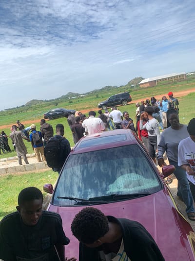 The image captures a group of people gathered outdoors in Gwafan, Nigeria. The scene appears to be informal, possibly a social gathering or a casual meeting. Several individuals are standing and sitting around a maroon-colored car, with some looking towards the camera. The ground is a mixture of grass and dirt, suggesting a rural or semi-rural setting. In the background, a large building with multiple windows and a light-colored roof is visible, potentially a school or an administrative building. The sky is partly cloudy, indicating daytime. The overall mood seems relaxed and communal, with people engaging with each other. There is no visible text in the image.
