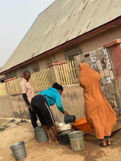Three individuals are depicted alongside a house exterior. A woman in an orange robe stands by a bucket. Another woman, wearing a blue jacket and black pants, fills a dark-colored bucket. A man in a light-colored shirt stands nearby. Several other buckets are stacked and scattered around the ground, some filled with water. The house features a metal roof, tiled exterior details, and barred windows.
