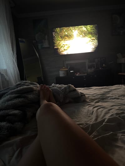 The image depicts a dimly lit bedroom scene, likely in Makiivka, Ukraine. The primary focus is a large television screen displaying a vibrant image of a waterfall in a lush, green environment. To the right of the television, a bed is visible, covered with crumpled grey bedding. A person's legs and feet are resting on a fluffy grey blanket draped over the bed. The rest of the room is cast in shadow, with hints of wall decorations and other objects on shelves behind the television, including what appears to be framed pictures. The overall mood is relaxed and cozy, suggesting the person is unwinding and watching television in bed. The time of day is not explicitly stated, but the darkness of the room and the illuminated screen imply it could be evening or nighttime. No weather conditions are discernible. The visible text on the television screen appears to be a digital clock displaying "20:44".