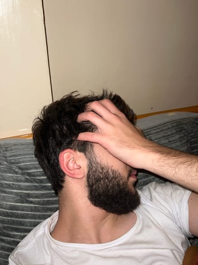 A close-up captures a person lying on a gray-striped surface, possibly a bed or couch. They are wearing a white t-shirt and have a dark beard and hair. One hand is resting on their head, fingers tangled in the hair. The individual's face is partially obscured. A light-colored wall is visible in the background, suggesting an indoor setting. The overall lighting appears dim, adding a sense of intimacy.