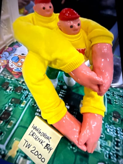 A close-up shot shows a pair of novelty doll hands wearing yellow long-sleeved shirts with ribbed cuffs. The hands are pressed together and appear to be part of a larger doll or toy that is mostly out of frame. The doll has two heads, both with red caps, rosy cheeks, and small black dots for eyes. One head is positioned to the left of the frame, while the other is slightly above and to the right of the first. The doll is resting on a surface that appears to be a circuit board with green and black components.

Tucked beneath one of the doll's arms is a small yellow sticky note with handwritten text in black marker. The text reads: "HANGOVER DRUNK BOY TW 2000".

The lighting in the image is bright and appears to be from an artificial source, possibly from a screen displaying the image. The background is dark, with some blurred blue and black shapes. The scene has a playful and potentially humorous tone, suggested by the novelty doll and the informal note. Given the location context of Taipei, Taiwan, this could be a souvenir or a unique item found in that region.