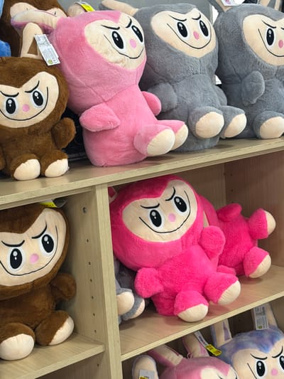 The image shows a shelf filled with stuffed animals, predominantly in shades of pink, brown, and gray. On the top shelf, from left to right, are a brown monkey-like stuffed animal, a large pink stuffed animal resembling a cartoon character with a hood, and two gray stuffed animals with similar facial features. The brown and pink stuffed animals are positioned at the front of the shelf. Below them, on the second shelf, is another large pink stuffed animal, lying down, with its face visible. To the right of this pink stuffed animal, a portion of a gray stuffed animal is visible. On the bottom shelf, partially visible, are more stuffed animals, including what appears to be a purple and white rabbit-like character. The shelves are made of light-colored wood and are part of a retail display. There are price tags and labels attached to some of the stuffed animals. The lighting suggests an indoor setting, likely a store. The image does not provide any specific information or visual cues to determine its location in Due Carrare, Italy.