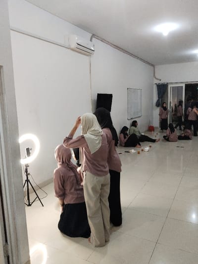 The image depicts a room with a tiled floor, white walls, and a ceiling with a light fixture. The room is located in the city of Telukjambe, Indonesia, based on coordinates -6.308 latitude and 107.282 longitude. There is an air conditioner mounted on the wall and a ring light set up on a tripod in the lower left corner of the room. Five women are in the room, all wearing hijabs. One woman is kneeling in the foreground, and the others are standing in a group in the center of the room. They are talking and laughing. There are also several people in the background, sitting or standing in a group, while others are walking out of the room. There are papers, cups, and a drink spilled on the floor. The scene suggests a casual, relaxed, and informal setting.