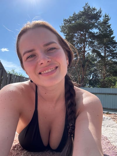 A close-up, low-angle selfie of an adult female is depicted, reportedly taken in Rusalevka, Russia. The woman has fair skin, light brown hair styled with a visible braid over her right shoulder, and a broad smile revealing her upper teeth. She is wearing a black bikini or halter-neck top, exposing her chest and shoulders, which show multiple small, dark spots. The background consists of a bright, clear blue sky with a strong sunflare in the upper-left quadrant and minimal cloud cover. Several large evergreen trees are visible in the mid-ground and background, predominantly on the right side. A light-colored fence runs horizontally across the mid-background on the right, and a section of a wooden fence is visible on the left. The woman is positioned on a patterned, textured surface, possibly a mat or blanket, with visible reddish-brown and lighter sections. The scene is brightly lit by direct natural sunlight.