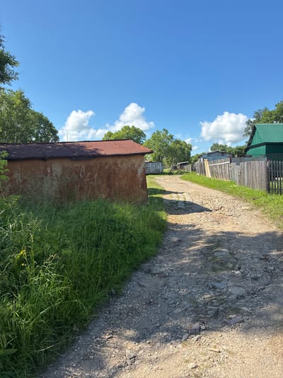 An outdoor, eye-level, medium shot captures a dirt road in Gornye Klyuchi, Russia, under a clear blue sky dotted with white, fluffy clouds. The road is flanked by overgrown green grass and various structures. On the left, a weathered, reddish-brown building with a rusty, corrugated metal roof stands prominently, partially obscured by tall weeds. To the right, a weathered wooden fence runs along the road, and beyond it, a green-roofed structure is visible. The road itself is unpaved, appearing rough and rocky in places, with shadows of trees dappling its surface. The scene suggests a rural or semi-rural setting within the town.