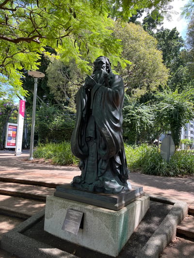The image displays a full-length, dark bronze statue of a figure, identifiable as Confucius, standing outdoors in a lush green park setting. The statue depicts an elderly male with a long beard, wearing elaborate flowing robes, and holding what appears to be a scroll or book. It stands on a grey stone pedestal, positioned beside a paved pathway or steps.

The background is dominated by dense, vibrant green foliage and numerous trees, suggesting a tropical or sub-tropical environment. Bright daylight illuminates the scene, casting clear shadows on the ground and the statue, indicating a sunny late morning or early afternoon.

A small, rectangular plaque is affixed to the front of the statue's pedestal. Although some text is small, discernible words include "THIS SCULPTURE," "CONFUCIUS," "REPLICA," "BRONZE," "GIFT," "BRISBANE," and "GUANGZHOU," along with "SISTER CITIES." This indicates the statue is a replica of Confucius, likely a gift exchanged between Brisbane and its sister city, Guangzhou.

To the left of the statue, a light pole is visible next to an informational sign. The sign prominently features "South Bank" in white text on a pink background, below which are several smaller pictorial panels and the word "JANUARY." The overall scene portrays a cultural landmark within a public park, located in South Brisbane, Australia.