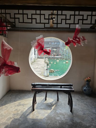 In a room with textured, concrete flooring and light-colored walls, two ornamental paper goldfish hang from the ceiling, suspended by thin threads. A dark wooden table with intricate carvings stands in the center of the room. On the table rests a guqin, a traditional Chinese stringed instrument. To the right of the table, a blue and white porcelain vase holds a small arrangement of light-colored flowers. A circular window dominates the background, offering a view of a serene pond with stepping stones and rocks. Beyond the pond, traditional Chinese architecture can be seen. The room is illuminated by sunlight casting long shadows on the floor. This scene appears to be located in Xiaoshan District, China.
