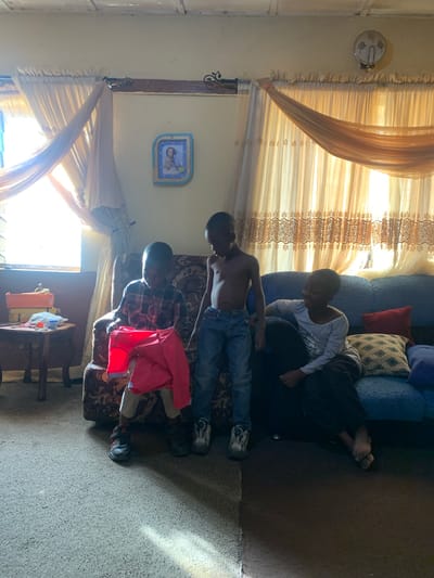 In Rafin Bauna, Nigeria, several individuals are gathered indoors. Two boys are visible on a floral patterned couch with a red jacket resting nearby. A third boy sits on a blue, pillowy couch. The setting features patterned curtains and walls decorated with what appears to be a clock. A window is visible in the background, adding to the indoor environment. The floor appears to be bare.