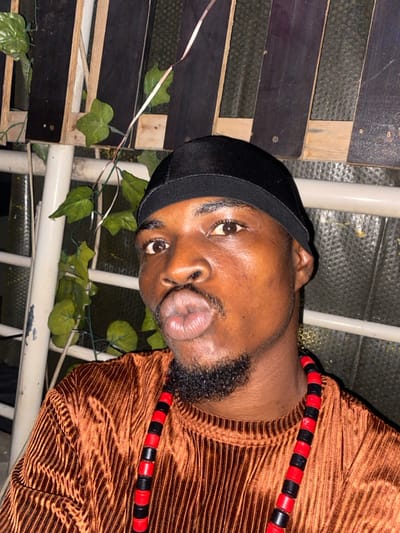A close-up shot shows a person wearing a black durag, a brown, textured, long-sleeved top, and a red and black beaded necklace. The person has dark skin, a beard, and is puckering their lips. Behind the person are plants with green leaves and a wooden pallet. White metal bars are also visible. The image is shot from a low angle and is slightly rotated to the side.