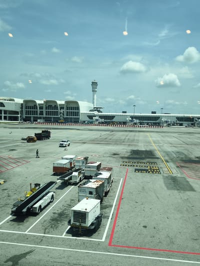 This image captures a scene at Kuala Lumpur International Airport (KLIA) in Sepang, Malaysia. Visible in the foreground are several pieces of airport ground support equipment parked within painted parking lanes. These include a baggage conveyor belt loader, multiple cargo containers, and an air conditioning unit.  A person is standing near a small loader.  

In the middle distance, more airport vehicles are present, including a white car and a truck. The airport terminal building and air traffic control tower are clearly visible in the background under a partly cloudy sky.  Airport signage and ground markings are evident throughout the image. Reflections from the interior are seen near the top of the photo.